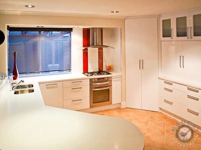 White gloss kitchen with a curved island bench, terracotta floor tiles and a red splashback accent