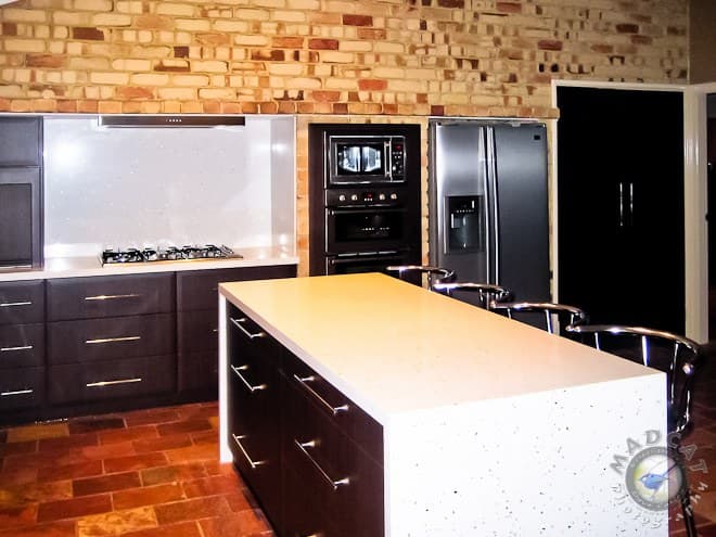 Completed kitchen renovation with a full-height feature brick wall, dark cabinetry, a waterfall stone island bench and integrated stainless fridge, Joondalup job